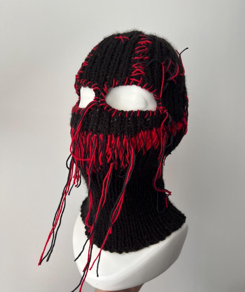 May include: Black knitted balaclava with red stitching and frayed edges. The balaclava has eye holes and covers the head and neck.
