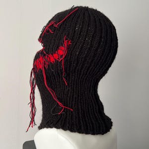 May include: Black knitted balaclava with red stitching detail. The balaclava has a hole for the eyes and mouth, and the stitching is visible around the edges.