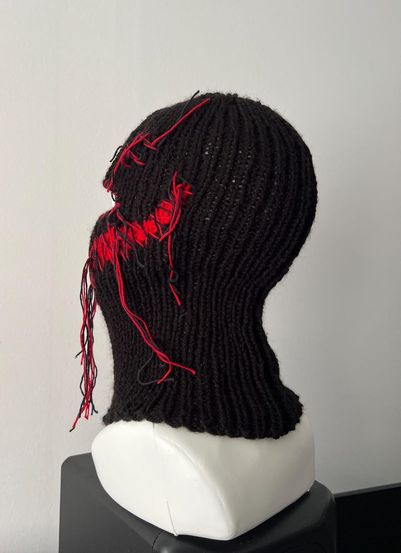 May include: Black knitted balaclava with red stitching detail. The balaclava has a hole for the eyes and mouth, and the stitching is visible around the edges.