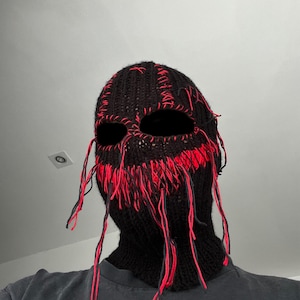 May include: A black knitted balaclava with red stitching and long strands of yarn hanging from the bottom. The balaclava has eye and mouth holes.