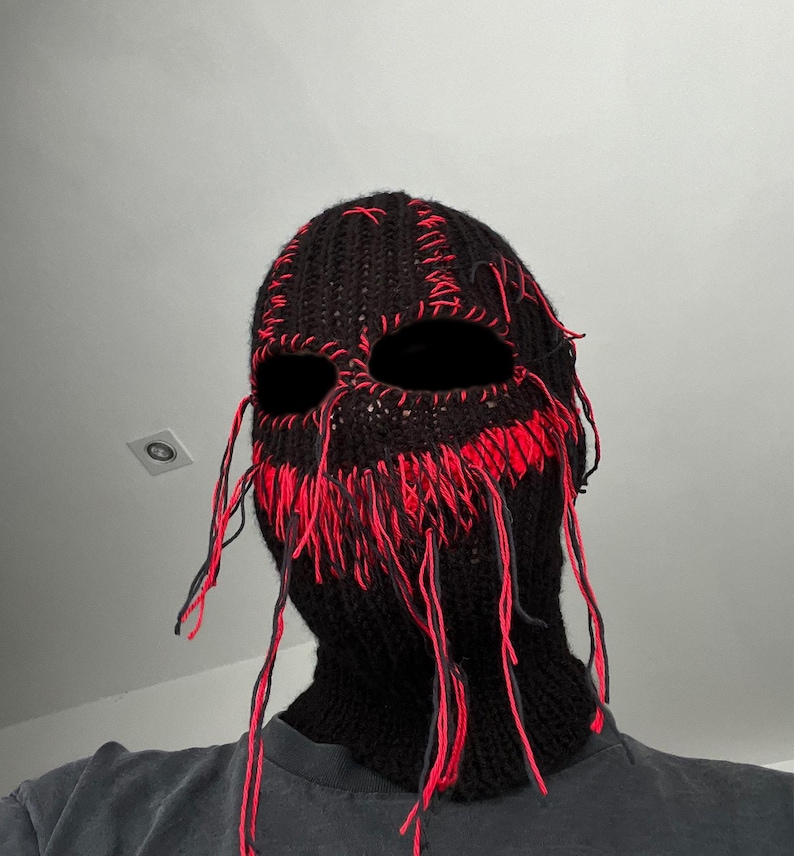 May include: A black knitted balaclava with red stitching and long strands of yarn hanging from the bottom. The balaclava has eye and mouth holes.