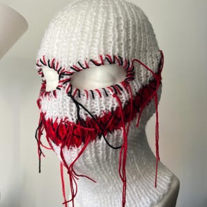 Puede incluir: Balaclava blanca de punto con costuras rojas y negras alrededor de los agujeros para los ojos. Las costuras están sueltas y cuelgan, creando un aspecto desordenado y desgastado.