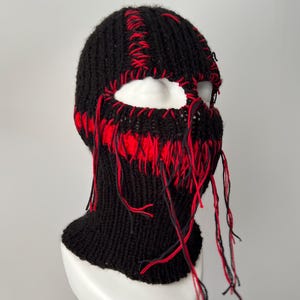 May include: Black knitted balaclava with red stitching and long red and black yarn strands hanging from the bottom.