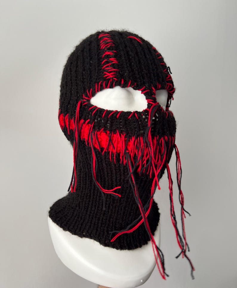 May include: Black knitted balaclava with red stitching and long red and black yarn strands hanging from the bottom.