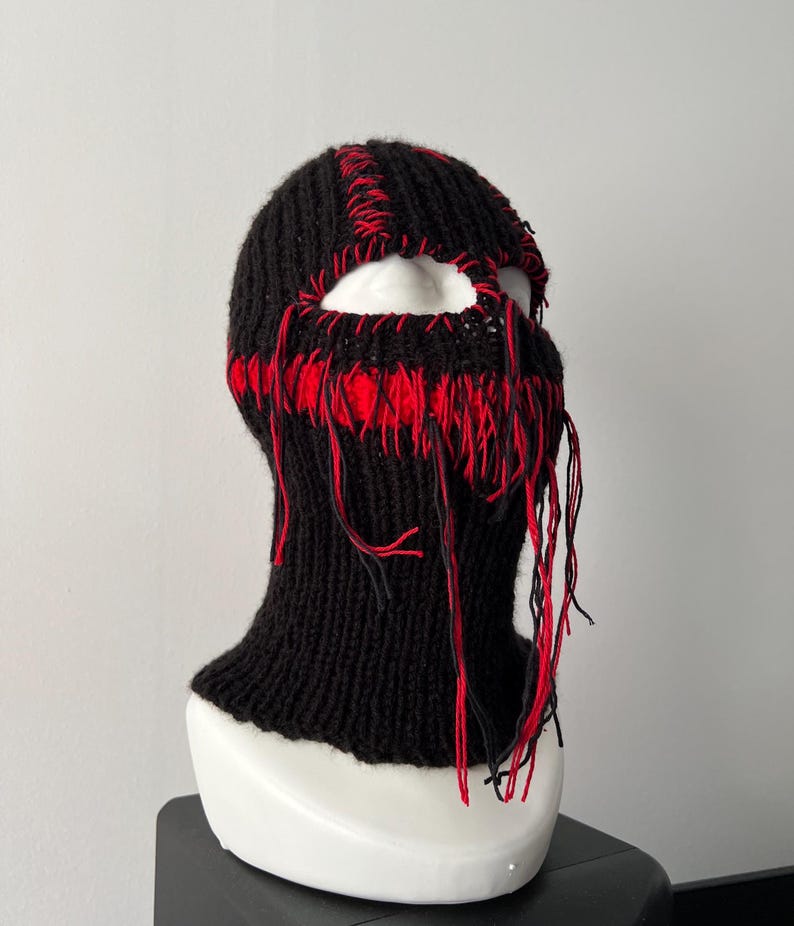 May include: Black knitted balaclava with red stitching and frayed edges. The balaclava has a hole for the eyes and nose.