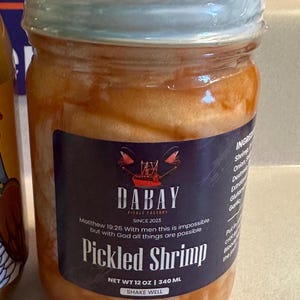 May include: A glass jar filled with pickled shrimp in a clear brine. The jar has a black label with the text "DABAY PICKLE FACTORY SINCE 2023" and "Pickled Shrimp". The label also includes the scripture verse "Matthew 19:26 With men this is impossible but with God all things are possible". The jar is 12 ounces or 340 milliliters.