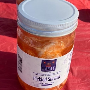 May include: A clear glass jar with a white lid and a label that reads "BABAY Pickled Shrimp." The jar contains shrimp in a reddish-orange brine. The net weight is 12 oz (340g).