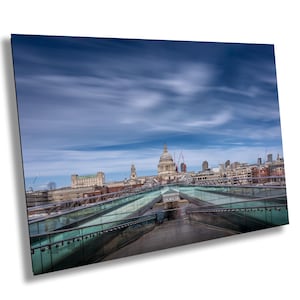 Puede incluir: Una vista de la Catedral de San Pablo en Londres, Inglaterra, desde un puente peatonal. El cielo es de un azul claro con nubes blancas. El puente está hecho de vidrio y metal.