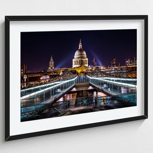 Puede incluir: Fotografía enmarcada de la Catedral de San Pablo en Londres por la noche. La cúpula iluminada y los edificios circundantes se reflejan en el cristal de una pasarela. El cielo es azul oscuro y la imagen está enmarcada en negro.