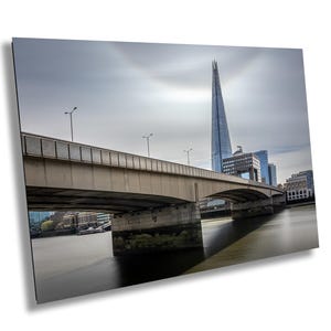 Op de afbeelding: Een grijze betonnen brug boogt over een rivier met een moderne wolkenkrabber op de achtergrond. De lucht is bewolkt en het water is kalm.