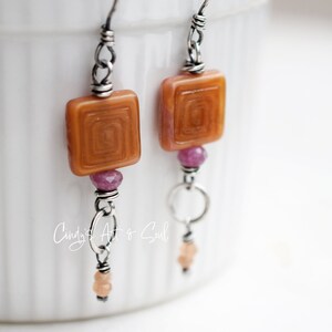 May include: Handmade earrings with square, amber-colored glass beads featuring a spiral design. The earrings also include pink and peach-colored beads, and silver-toned metal accents. The earrings are suspended from silver-toned ear wires.