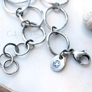 K&ouml;nnte beinhalten: Ein silberfarbenes Kettenarmband mit miteinander verbundenen kreisf&ouml;rmigen Gliedern in dunkler, oxidierter Optik. Das Armband verf&uuml;gt &uuml;ber einen Karabinerverschluss und einen kleinen, runden Metallanh&auml;nger mit eingraviertem f&uuml;nfzackigen Stern.