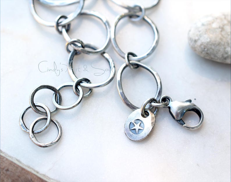 K&ouml;nnte beinhalten: Ein silberfarbenes Kettenarmband mit miteinander verbundenen kreisf&ouml;rmigen Gliedern in dunkler, oxidierter Optik. Das Armband verf&uuml;gt &uuml;ber einen Karabinerverschluss und einen kleinen, runden Metallanh&auml;nger mit eingraviertem f&uuml;nfzackigen Stern.