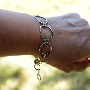 K&ouml;nnte beinhalten: Ein silberfarbenes Armband, das aus mehreren miteinander verbundenen, unregelm&auml;&szlig;ig geformten kreisf&ouml;rmigen Gliedern besteht. Die Kette zeigt eine Reihe von unterschiedlich gro&szlig;en, offenen Ringen, die miteinander verbunden sind, mit einem kleinen Anh&auml;nger am Ende.