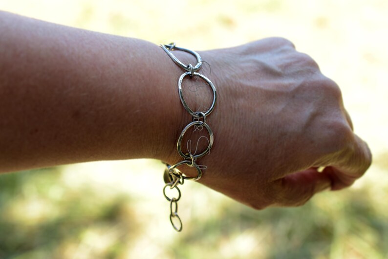 K&ouml;nnte beinhalten: Ein silberfarbenes Armband, das aus mehreren miteinander verbundenen, unregelm&auml;&szlig;ig geformten kreisf&ouml;rmigen Gliedern besteht. Die Kette zeigt eine Reihe von unterschiedlich gro&szlig;en, offenen Ringen, die miteinander verbunden sind, mit einem kleinen Anh&auml;nger am Ende.