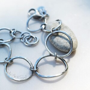 K&ouml;nnte beinhalten: Ein silberfarbenes Kettenarmband, das aus mehreren unregelm&auml;&szlig;igen, geh&auml;mmerten kreisf&ouml;rmigen Gliedern unterschiedlicher Gr&ouml;&szlig;e besteht. Das Armband verf&uuml;gt &uuml;ber einen Karabinerverschluss und einen kleinen runden Anh&auml;nger in der N&auml;he des Verschlusses.