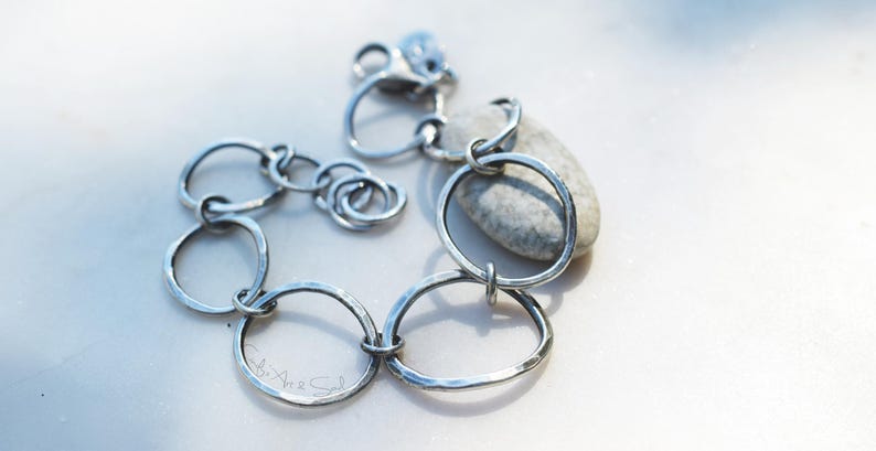 K&ouml;nnte beinhalten: Ein silberfarbenes Kettenarmband, das aus mehreren unregelm&auml;&szlig;igen, geh&auml;mmerten kreisf&ouml;rmigen Gliedern unterschiedlicher Gr&ouml;&szlig;e besteht. Das Armband verf&uuml;gt &uuml;ber einen Karabinerverschluss und einen kleinen runden Anh&auml;nger in der N&auml;he des Verschlusses.