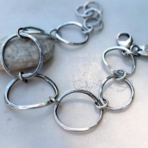 K&ouml;nnte beinhalten: Ein silberfarbenes Kettenarmband bestehend aus mehreren unregelm&auml;&szlig;igen, geh&auml;mmerten kreisf&ouml;rmigen Gliedern. Das Armband verf&uuml;gt &uuml;ber einen Karabinerverschluss und einen kleinen, ovalen Metallanh&auml;nger mit eingraviertem Symbol. Der Text Cindy's Art & Soul ist sichtbar.