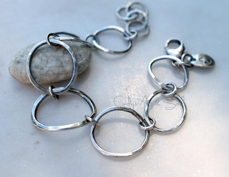 K&ouml;nnte beinhalten: Ein silberfarbenes Kettenarmband bestehend aus mehreren unregelm&auml;&szlig;igen, geh&auml;mmerten kreisf&ouml;rmigen Gliedern. Das Armband verf&uuml;gt &uuml;ber einen Karabinerverschluss und einen kleinen, ovalen Metallanh&auml;nger mit eingraviertem Symbol. Der Text Cindy's Art & Soul ist sichtbar.
