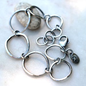 K&ouml;nnte beinhalten: Ein silberfarbenes Armband aus mehreren miteinander verbundenen, unregelm&auml;&szlig;igen kreisf&ouml;rmigen Gliedern mit geh&auml;mmertem Finish. Das Schmuckst&uuml;ck verf&uuml;gt &uuml;ber einen Karabinerverschluss und einen kleinen, runden Anh&auml;nger mit eingepr&auml;gtem Stern.