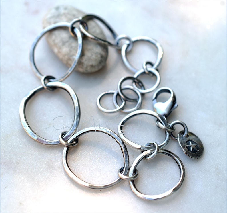 K&ouml;nnte beinhalten: Ein silberfarbenes Armband aus mehreren miteinander verbundenen, unregelm&auml;&szlig;igen kreisf&ouml;rmigen Gliedern mit geh&auml;mmertem Finish. Das Schmuckst&uuml;ck verf&uuml;gt &uuml;ber einen Karabinerverschluss und einen kleinen, runden Anh&auml;nger mit eingepr&auml;gtem Stern.