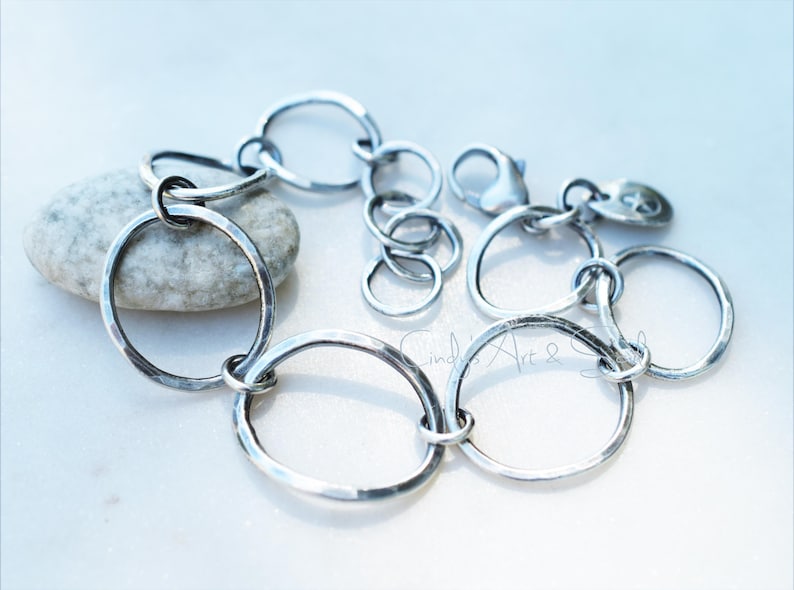 K&ouml;nnte beinhalten: Ein silberfarbenes Kettenarmband bestehend aus mehreren miteinander verbundenen, geh&auml;mmerten kreisf&ouml;rmigen Gliedern unterschiedlicher Gr&ouml;&szlig;e. Das Armband verf&uuml;gt &uuml;ber einen Karabinerverschluss und eine Verl&auml;ngerungskette mit einem kleinen runden Anh&auml;nger.