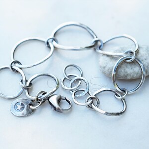 K&ouml;nnte beinhalten: Ein silberfarbenes Kettenarmband aus unregelm&auml;&szlig;igen, handgeh&auml;mmerten kreisf&ouml;rmigen Gliedern. Das Armband verf&uuml;gt &uuml;ber einen Karabinerverschluss und einen kleinen, runden Anh&auml;nger mit einem eingepr&auml;gten f&uuml;nfzackigen Stern. Das Metall hat ein dunkles, oxidiertes Finish.