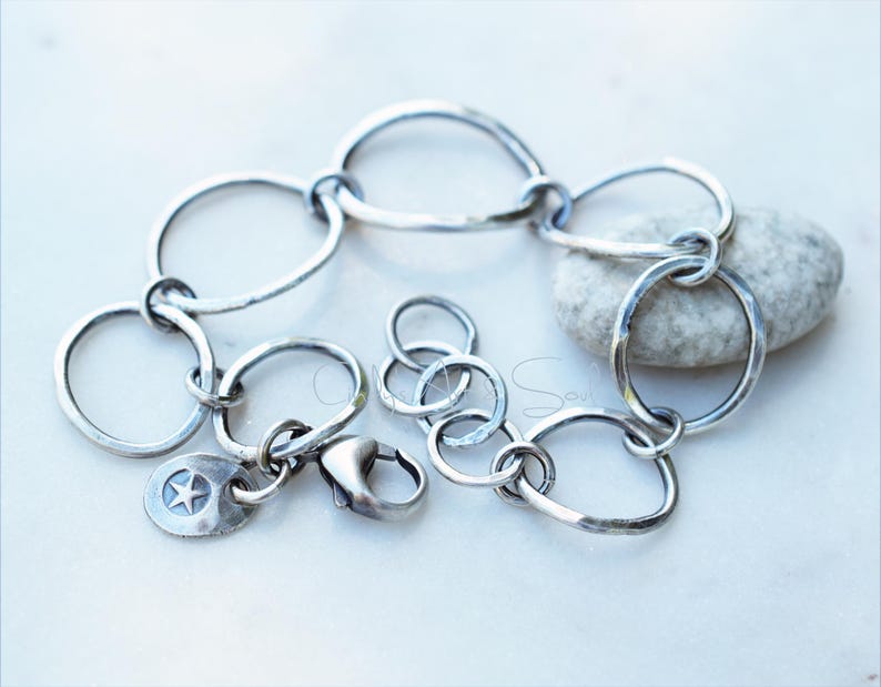 K&ouml;nnte beinhalten: Ein silberfarbenes Kettenarmband aus unregelm&auml;&szlig;igen, handgeh&auml;mmerten kreisf&ouml;rmigen Gliedern. Das Armband verf&uuml;gt &uuml;ber einen Karabinerverschluss und einen kleinen, runden Anh&auml;nger mit einem eingepr&auml;gten f&uuml;nfzackigen Stern. Das Metall hat ein dunkles, oxidiertes Finish.