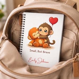 Puede incluir: Un cuaderno de espiral con una ilustración de dibujos animados de dos monos abrazándose. Uno es naranja, el otro marrón, con un globo rojo en forma de corazón. El texto "Small, But Brave" y un nombre están impresos en la portada. El cuaderno está dentro de una bolsa beige.