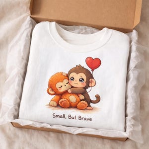 May include: White crew neck t-shirt with a cartoon illustration of two hugging monkeys, one brown and one orange, and a red heart balloon. The text "Small, But Brave" is printed below the image. The shirt is presented in a cardboard box.