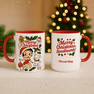 Op de afbeelding: Twee witte keramische mokken met rode binnenkant en handvatten. Eén mok heeft Betty Boop in een kerstmuts met de woorden "Merry Christmas". De andere mok zegt "Merry Christmas, Sweethearts!" met de naam "Olivya's Mug".