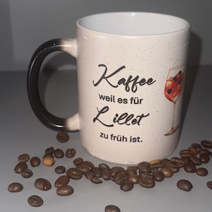 Könnte beinhalten: Weiße Keramik-Kaffeetasse mit schwarzem Henkel und schwarzem Text "Kaffee weil es für Lillet zu früh ist." Die Tasse hat eine glitzernde Oberfläche und eine bunte Illustration eines Weinglases mit Beeren.
