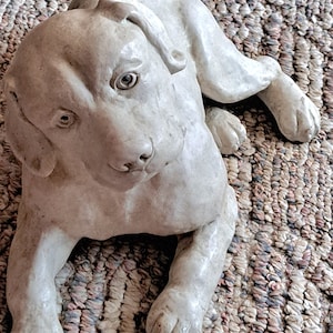 May include: A light-colored, ceramic dog figurine in a relaxed pose. The dog is lying down with its head turned, and its paws are extended. The figurine is on a textured, multi-colored rug.