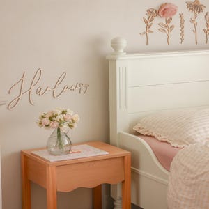 May include: A wooden wall cutout featuring the name Harlow in a cursive script font, positioned above a light wood nightstand. The nightstand holds a clear glass vase filled with a bouquet of small pink and white artificial roses.