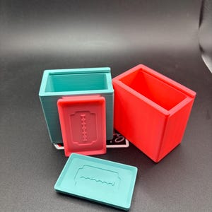 Op de afbeelding: Twee rechthoekige 3D-geprinte opbergbakjes met twee bijpassende deksels. Eén bakje en deksel zijn lichtblauw, terwijl het andere bakje en deksel felrood zijn. Elk deksel is voorzien van een reliëf van een dubbelzijdig scheermesje.