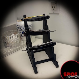 May include: A black wooden stand with three tiers for holding straight razors. The stand is holding three razors, one with a gold handle and two with black handles.