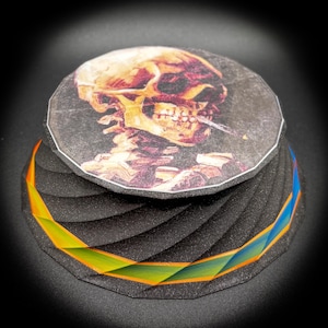 Op de afbeelding: Een decoratief, meerlagig object met een schedelafbeelding. De schedel is afgebeeld in bruine en gouden tinten, met een sigaret. De basis is zwart met een geometrisch ontwerp en een band van oranje, groen en blauw.