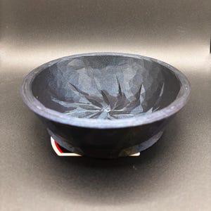 Peut inclure: Un bol en bois sculpté bleu foncé avec une surface texturée. Le bol a une forme large et peu profonde et un motif géométrique sculpté à l'intérieur. Le bol repose sur une petite base décorative.