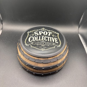 Può includere: Un contenitore rotondo a forma di barile con una texture color legno marrone e fasce nere in stile metallico con piccoli rivetti. Il coperchio nero presenta un bordo decorativo color oro e il testo "SPOT COLLECTIVE ARTISAN GOODS EST. 2024".
