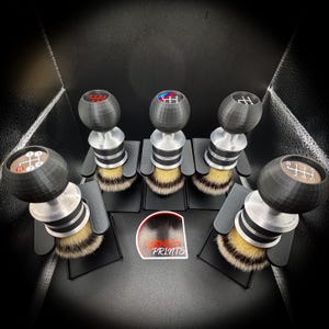 Op de afbeelding: Vijf zwarte en zilveren scheerkwasten met versnellingspookknopontwerpen. Elke borstel heeft een ander schakelpatroon in rood, blauw en bruin. De borstels zijn gerangschikt op een zwart oppervlak.