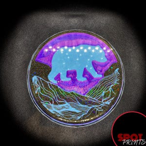 May include: A round, clear resin coaster with a purple and blue design. The design features a silhouette of a bear walking across a mountain range. The coaster has a glittery finish.