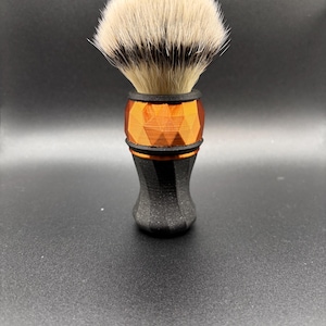 Puede incluir: Una brocha de afeitar con una cabeza de cerdas clara y completa y un mango geométrico naranja y negro. El cepillo tiene una banda negra en la base de las cerdas y otra en la parte superior del mango. El mango tiene un diseño facetado.