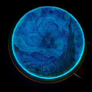 May include: A round, glowing art piece featuring a depiction of Van Gogh's "Starry Night." The artwork is rendered in shades of blue and black, with a bright, neon-blue border. The image showcases the swirling sky, village, and cypress tree.