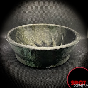 May include: A black and green ceramic bowl with a textured, speckled surface. The bowl is round and has a wide, shallow opening.