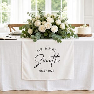 May include: A white fabric table runner featuring the black printed text MR. & MRS. Smith and the date 06.27.2026. The runner is draped over a white tablecloth on a table decorated with a large floral centerpiece, a small cake, and two candles.