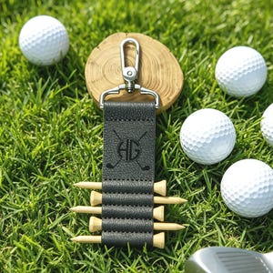 May include: A dark gray textured leather golf tee holder keychain with a silver metal lobster clasp. The holder features a circular monogram with the letters HG and crossed golf clubs, containing five light-colored wooden golf tees.