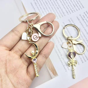 Peut inclure: Porte-clés dorés avec des breloques. L'un présente un croissant de lune avec une perle et une poignée ornée de cristaux. L'autre a un cœur et un nœud. Autres breloques : un cœur et une spirale.