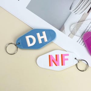 K&ouml;nnte beinhalten: Drei Schl&uuml;sselanh&auml;nger im Retro-Stil. Einer ist blau mit "DH" in Wei&szlig;, ein anderer ist wei&szlig; mit "NF" in Pink und Orange, und der dritte ist lila mit "MP" in Pink. Jeder Schl&uuml;sselanh&auml;nger hat einen silbernen Ring.