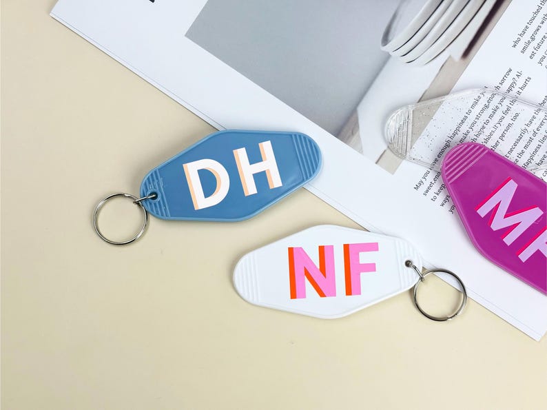 K&ouml;nnte beinhalten: Drei Schl&uuml;sselanh&auml;nger im Retro-Stil. Einer ist blau mit "DH" in Wei&szlig;, ein anderer ist wei&szlig; mit "NF" in Pink und Orange, und der dritte ist lila mit "MP" in Pink. Jeder Schl&uuml;sselanh&auml;nger hat einen silbernen Ring.