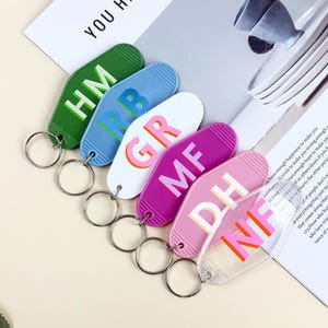 K&ouml;nnte beinhalten: Eine Sammlung farbenfroher Schl&uuml;sselanh&auml;nger in Gr&uuml;n, Blau, Wei&szlig;, Lila, Rosa und Transparent. Jeder Schl&uuml;sselanh&auml;nger hat ein einzigartiges Design mit zwei Buchstaben in kr&auml;ftigen, kontrastierenden Farben. Die Schl&uuml;sselanh&auml;nger sind an silbernen Schl&uuml;sselringen befestigt.